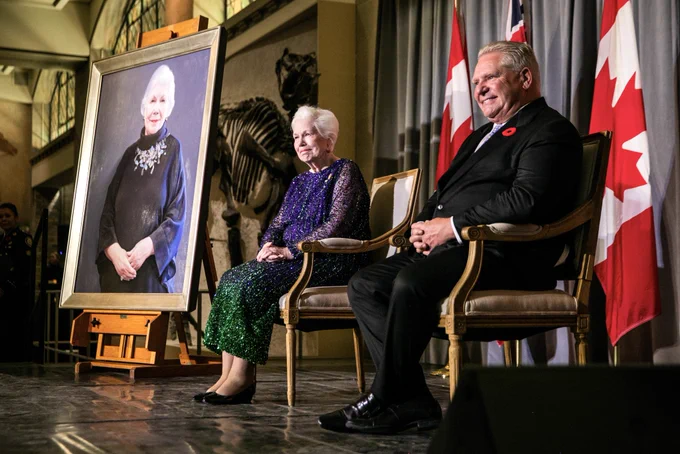 The Lieutenant Governor and Premier sat in chairs on a stage, with Canadian flags behind them. To their right is a portrait of the Lieutenant Governor