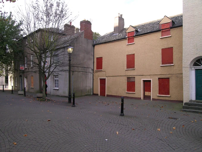 A photograph of two houses in Cathedral Square, Waterford, from 2005 prior to their conservation and repair under the Built Heritage Investment Scheme and Historic Structures Fund.  The houses are vacant, in poor repair, with the house on the left showing inappropriate uPVC windows and a cement rendered surface finish and the house on the right, set back slightly, showing boarded up fittings.  There is extensive cobble locking in the foreground