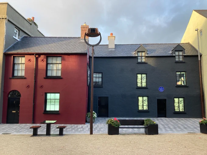 A photograph of two houses in Cathedral Square, Waterford, from November 2023 following their conservation under the Built Heritage Investment Scheme and Historic Structures Fund.  The houses are in good repair and have had historically-correct timber sash and timber casement windows inserted: the windows are illuminated with the branding of the Irish Wake Museum.  The roofs of both houses have been restored with natural slate.  The house on the left has been repainted in a rose red while the house on the right has been repainted in a dark blue.   The foreground has been relandscaped with stone flags and a sandy-coloured gravel.