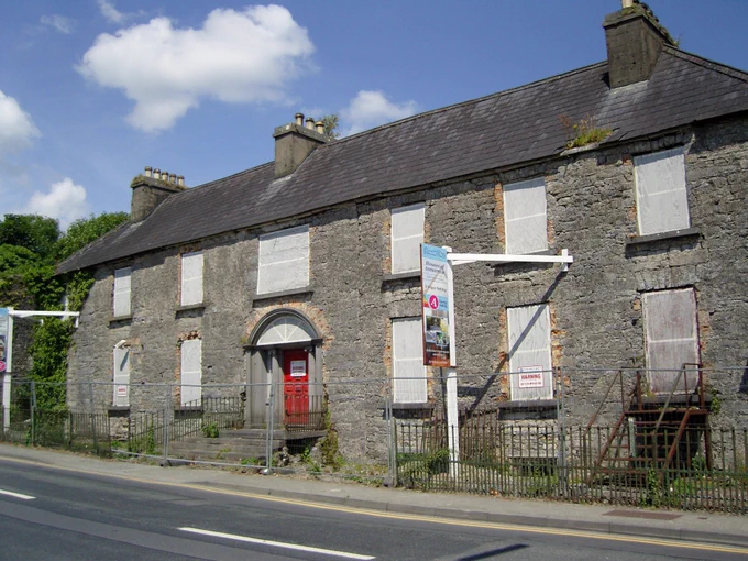 A photograph of Robe Villa, Ballinrobe, Mayo, from 2011 showing the house in poor repair.  The house is seven windows wide, two storeys high, and has a Classical doorcase of limestone at its centre.  All of the openings are boarded up.  The render has come away from the walls to reveal a rubble limestone construction.  A crack or masonry break to the right of the door is evidence that the house was originally much smaller but was extended at a later date.