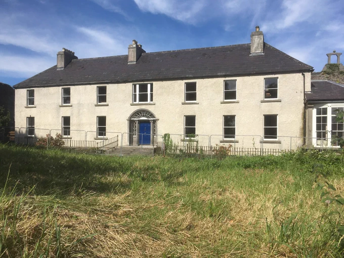 A photograph of Robe Villa, Ballinrobe, Mayo, from 2022 showing the house in good repair following its conservation under the Built Heritage Investment Scheme.  The Classical doorcase of limestone at its centre has been restored and shows a pretty lace-like fanlight.  All of the openings have been refitted with white-coloured timber sash windows.  The walls have been replastered with a creamy-coloured lime render.