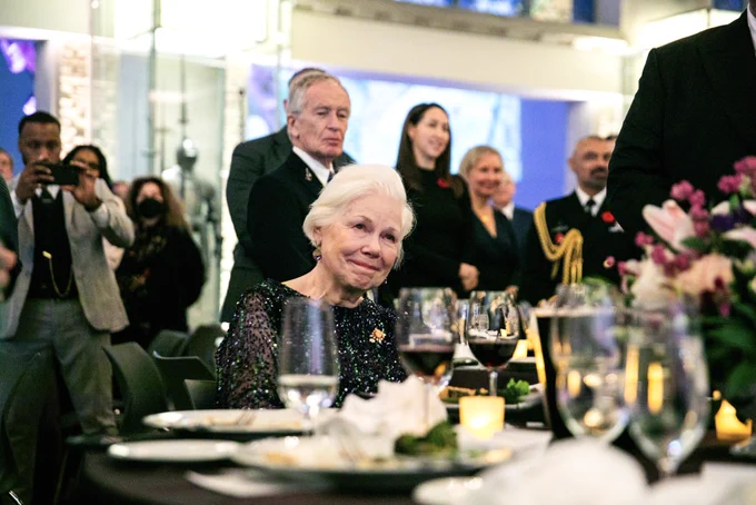 The Lieutenant Governor seated at a dining table, looking tearfully towards a stage out of the image frame
