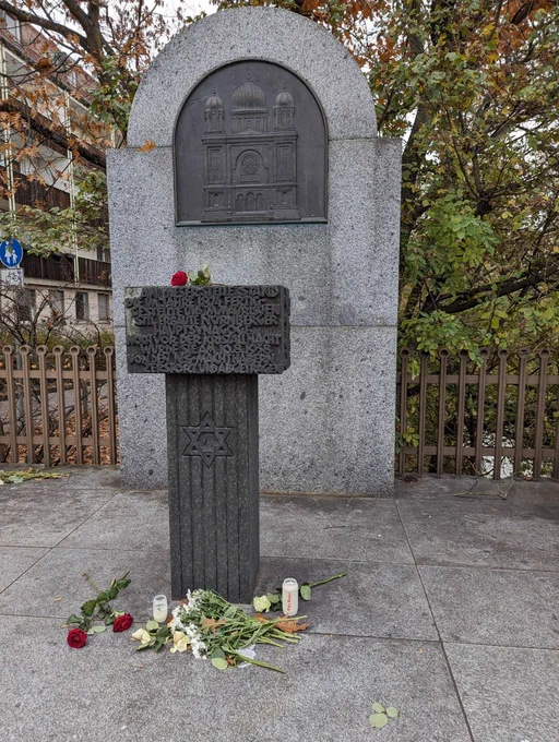 Herbstliches Bild des Denkmal der 1874 fertiggestellten Nürnberger Hauptsynagoge die am 10.8.1938 von den NS Machthabern zerstört würde. Im Hintergrund eine Marmorsteinwand mit Bronzerelief der Synagoge, davor ein Altar, mit Aufschrift über das Denkmal auf der Säule die den Altar trägt ist ein Davidsstern. Auf dem Altar liegt eine rote Rose. Davor liefen mehrere Blumen und zwei Grablichter, eines davon beschriftet in rot mit "#NieWieder".