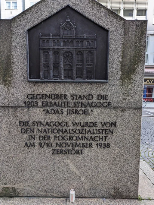 Denkmal zur Adas Jisroel Synagoge in Nürnberg. Eine Steintafel mit einem Bronze Relief der Synagoge, darauf Bronze Schrift "Gegenüber stand die 1903 erbaute Synagoge "Adas Jisroel"
Die Synagoge wurde von den Nationalsozialisten in der Pogromnacht am 9./10. November 1938 zerstört"
Darunter steht ein brennendes weißes Grablicht mit roter Hand geschriebener Aufschrift "#NieWieder"