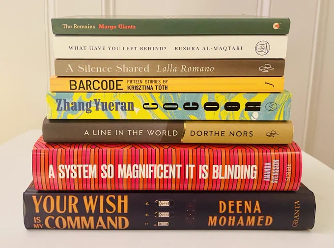 A photograph of the 8 titles shortlisted for the Warwick Prize for Women in Translation in 2023. The books are stacked on their side on a white table against a white background. The shortlisted titles are:
Dorthe Nors, A Line in the World, translated from Danish (Denmark) by Caroline Waight (Pushkin Press).
Lalla Romano, A Silence Shared, translated from Italian (Italy) by Brian Robert Moore (Pushkin Press).
Amanda Svensson, A System So Magnificent It Is Blinding, translated from Swedish (Sweden) by Nichola Smalley (Scribe UK).
Krisztina Tóth, Barcode, translated from Hungarian (Hungary) by Peter Sherwood (Jantar).
Zhang Yueran, Cocoon, translated from Chinese (China) by Jeremy Tiang (World Editions).
Margo Glantz, The Remains, translated from Spanish (Mexico) by Ellen Jones (Charco Press).
Bushra al-Maqtari, What Have You Left Behind? translated from Arabic (Yemen) by Sawad Hussain (Fitzcarraldo Editions).
Deena Mohamed, Your Wish Is My Command, translated from Arabic (Egypt).