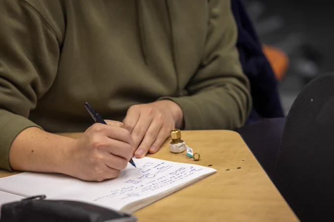 A close-up of a student taking notes in a notebook. Next to the notebook are two pieces of hardware.