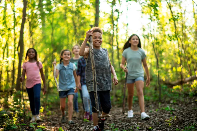a group of young children take a hike in the woods on a sunny spring day. They are each dressed casually and some have wooden walking sticks