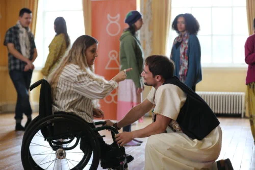A woman seated in a wheelchair speaks to a man kneeling to the front of her. He appears to listen intently. In the background are several onlookers.