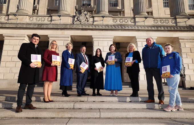 Image shows 9 people standing on steps at Stormont parliament buildings, each holding a report booklet