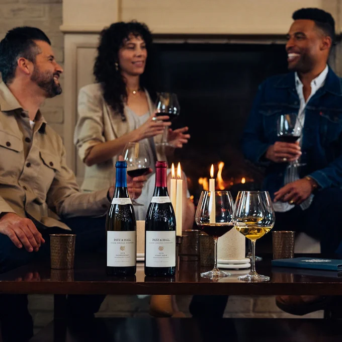 Three people are sitting by a fireplace, smiling and holding wine glasses. Bottle at Patz & Hall Pinot Noir and Chardonnay sits on the table. 