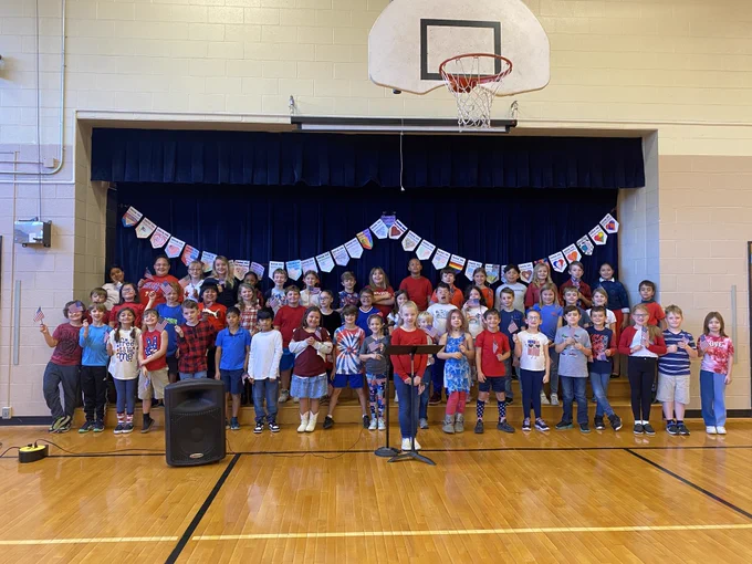 third graders stand in a group in front of the stage and hold american flags. they are getting ready to perform and they are all wearing red, white, and blue