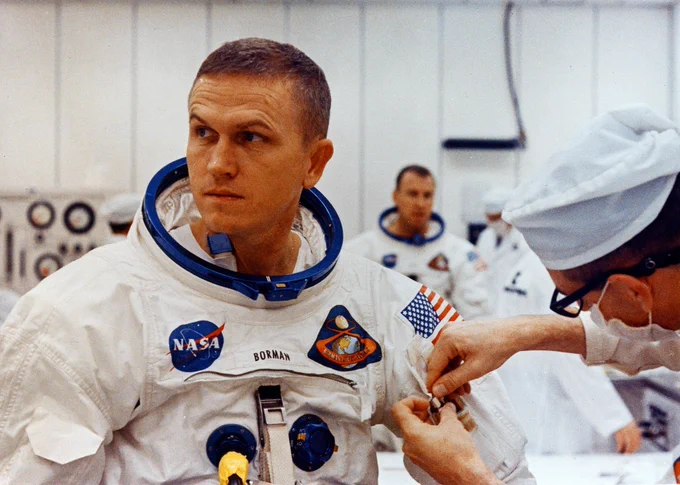 Frank Borman in white Apollo spacesuit looking toward the left of the frame. A spacesuit technician is adjusting something on his suit.