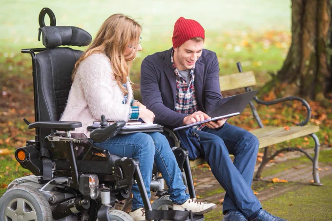 Photo of two people sitting outside, looking at a tablet. One is in an electric wheelchair.