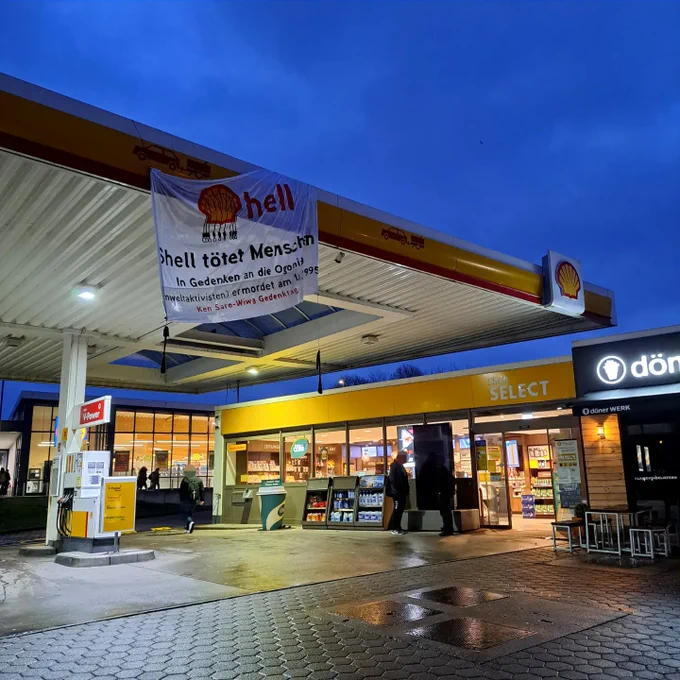Das Foto zeigt eine Shell-Tankstelle in der Abenddämmerung. Der Himmel ist blau bis lila gefärbt, was auf die blaue Stunde hindeutet. Über den Zapfsäulen hängt ein großes weißes Transparent mit der Aufschrift: "Shell tötet Menschen". Zusätzlicher Text auf dem Banner erinnert an die ermordeten Ogoni-Aktivisten von 1995 und bezieht sich auf das Ken-Saro-Wiwa-Gedenktages. Die Beleuchtung der Tankstelle ist eingeschaltet, was auf den Abend oder frühen Morgen schließen lässt. Kunden sind am Eingang des Shops zu sehen, und die Umgebung wirkt ruhig. Das Shell-Firmenlogo ist deutlich sichtbar, was im Kontrast zu der kritischen Botschaft des Banners steht. Die Szene ist friedlich, doch die Nachricht vermittelt eine ernste Botschaft über die Aktivitäten des Unternehmens und deren Auswirkungen.