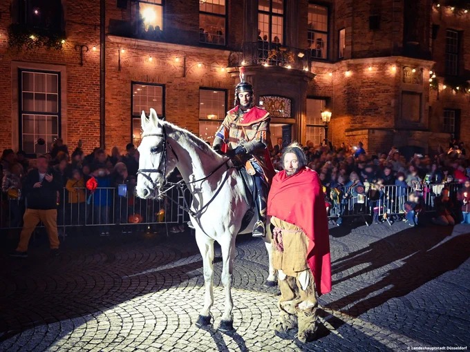 Sankt Martin und ein Bettler stehen vor dem Rathaus in Düsseldorf. Es ist bereits dunkel und das Rathaus ist stimmungsvoll beleuchtet. Sankt Martin sitzt auf einem Schimmel, der Bettler steht in einen roten Mantel gehüllt neben dem Pferd. Hinter den beiden sind unzählige Kinder und Erwachsene zu sehen, die sich den Umzug hinter einer Absperrung gespannt anschauen. Viele Kinder halten bunte Laternen in den Händen.