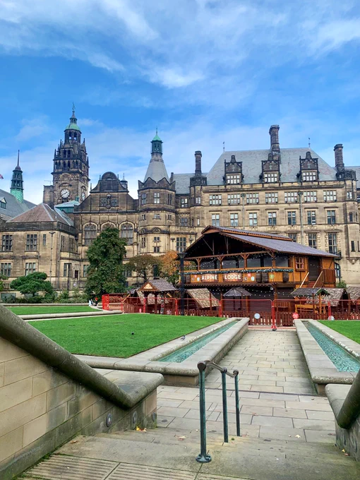 Sheffield Peace Gardens viewed from one of the entrance steps, showing an Alpine Lodge under construction in the centre of gardens with Sheffield Town Hall clock tower and building in the background, plus blue skies above.