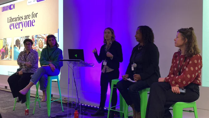 Five members of the LibraryOn team, who work for the British library sitting on green stalls at the front of a conference room in a panel, with one member of the team in the middle standing and presenting to the room.