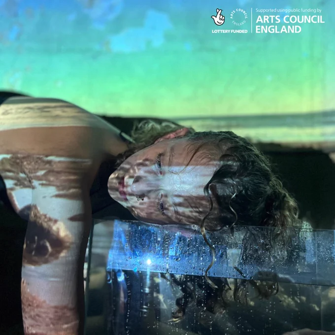 A white woman with blond curly hair rests her head on a tank full of water. She looks to the left of the camera. The image of a coast and shoreline is being projected onto her