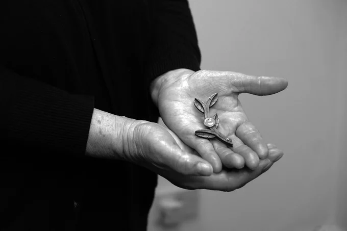 a black and white photograph of a pair of hands, one resting in the other, palm up, holding an olive branch shaped piece of walnut & enamel jewellery. 
