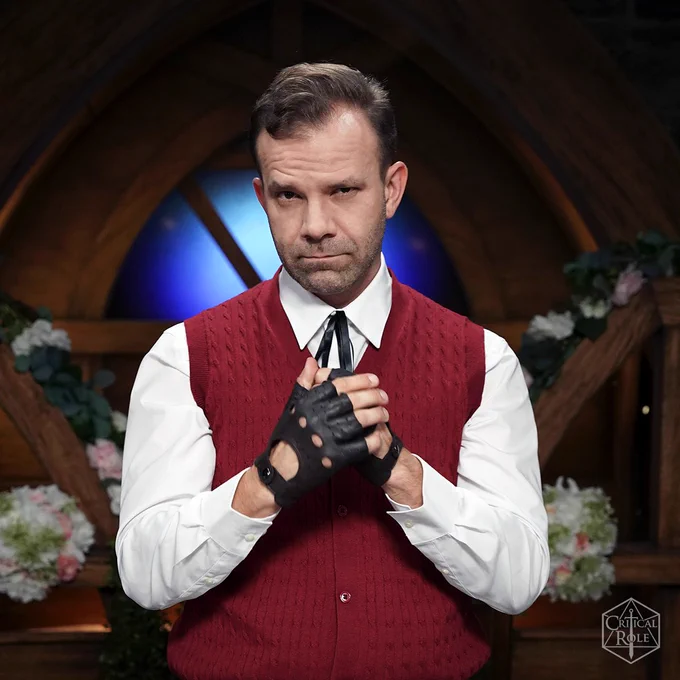 Liam O'Brien stands looking pensively in a red vest, high collared shirt, fingerless leather driving gloves, and a black bolo tie on the flower filled Critical Role set in this photo