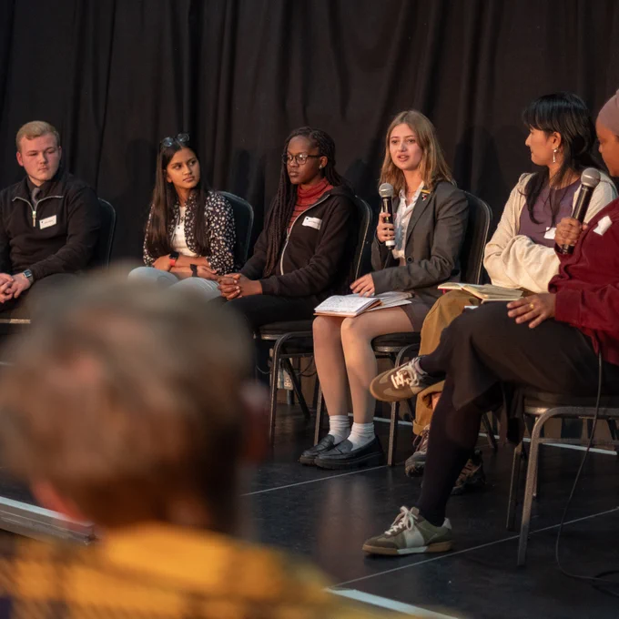 A panel of four young people sit on chairs on a stage, while one of them talks into a microphone to an audience. 
