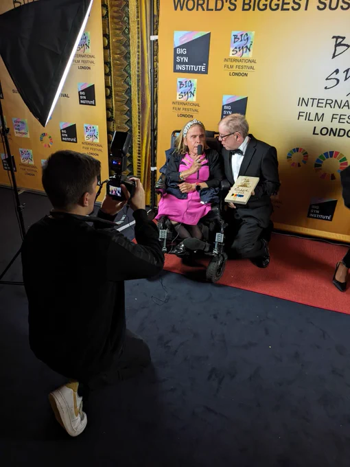 MLMC members Jackie and Paul hold a microphone as they give a media interview at the Big Syn film festival. Paul holds the award.