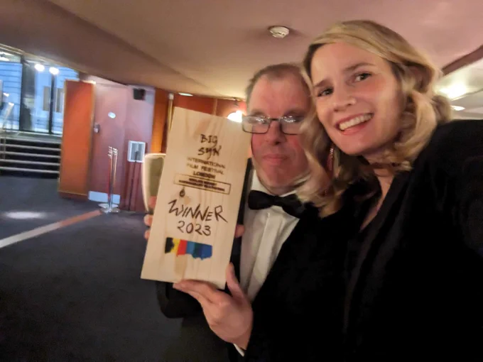 Paul and Grace pose with the Big Syn charity film of the year award in a lobby.