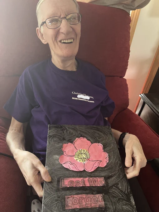 A man wearing a purple t-shirt holds up his artwork of a red poppy painted on a black background. The painting also says "lest we forget." 