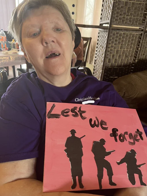 A woman wearing a purple t-shirt holds up her painted artwork. The canvas has a red background with silhouettes of soldiers and the words  "lest we forget"