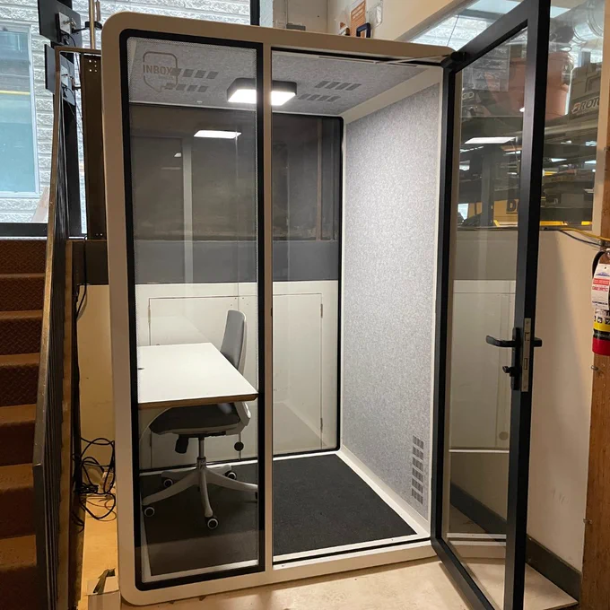 A ONE plus phone booth pod, featuring a desk, rolling chair, and light and ventilation system, covered by a tempered glass door. This phone booth is located by the staircase at the back of CSI Spadina's ground floor.