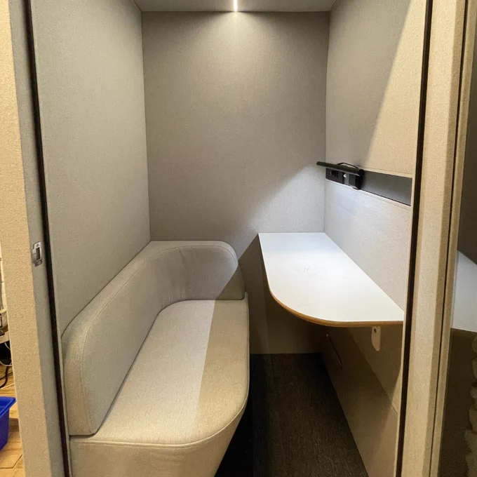 The inside of a TUESDAY model phone booth from Inbox Booths, featuring a cushioned bench and a table and small lamp attached to the wall.