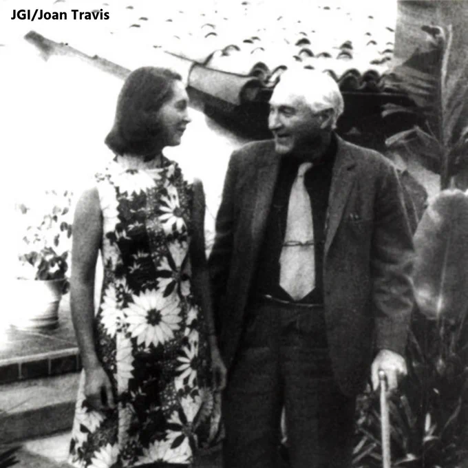 Black and white photo of an elderly man wearing a jacket and tie and walking with a cane (Dr. Louis Leakey) standing next to and speaking with a young woman in a floral dress (Dr. Jane Goodall). Photo credit: JGI/Joan Travis.