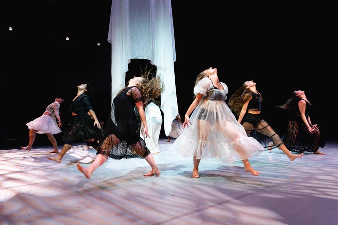 The image shows a group of actors and dancers on stage. They are dancing around and under a white hanging sheet. Four performers are wearing black undergarments visible through their sheer white clothes. The remaining four performers are in all black and lace. The pattern of clothing is one sheer white person after two black lace people. Photo by Manoo Sirivelu.