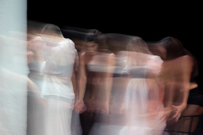 The image shows a group of dancers out of focus and in a blur as they perform. All the performers are wearing black undergarments visible through their sheer white clothes, except for the performer in the center, who is in all black and lace. Photo by Manoo Sirivelu.