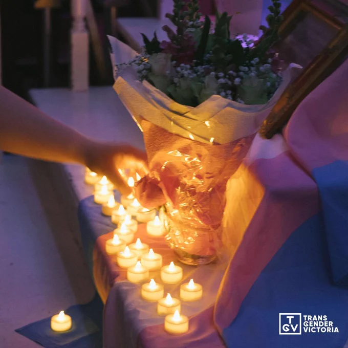  A photograph taken at a previous trans day of remembrance vigil. Photographed is a bouquet of flowers on series of steps that has been decorated with a trans flag. The flower bouquet is standing up in a jar and is surrounded by candles. On the left side of the frame is a hand incoming towards the flowers, placing down a flower.