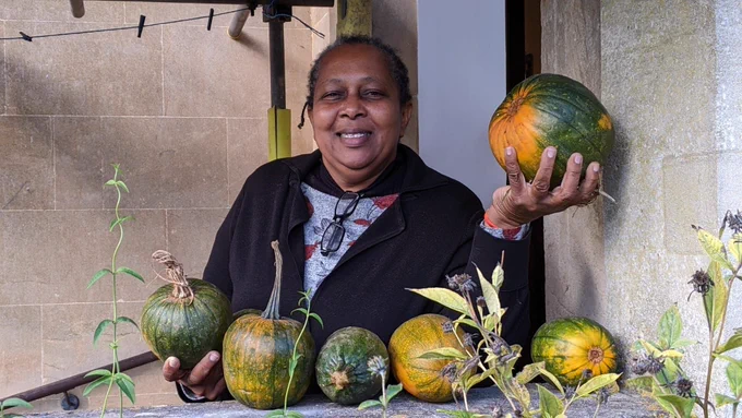 A woman smiling and holding two donated pumpkins, with a further four pumpkins on a wall.