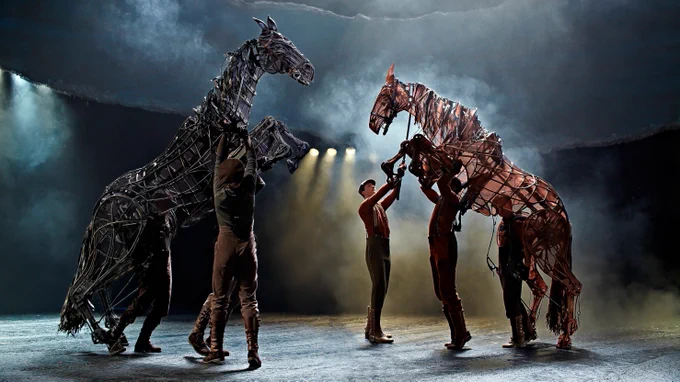 Two large puppet horses are moved by men using metal rods. The horses are positioned on their hind legs as though in battle and scared. Around them is mist.