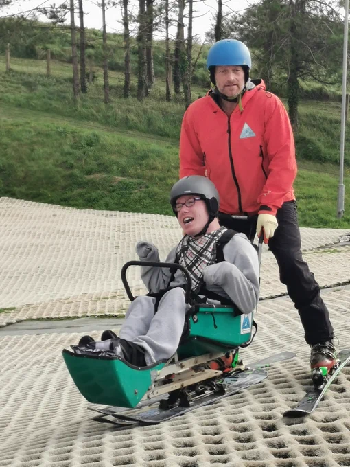 Arwel, wearing a grey outfit, is sitting on an accessible toboggan going gently down a dry slope. There is a ski instructor wearing a red jacket, with skis on his feet, pushing him along. They are both wearing helmets and are smiling happily. 