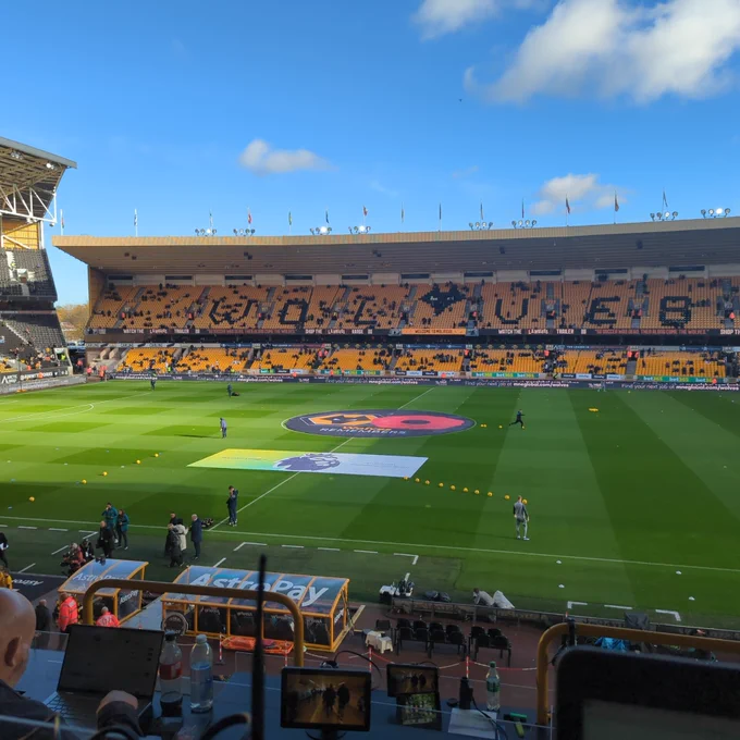 Photo of the Steve bull stand at Molineux slowly starting to fill up. A large circular banner covers the centre circle with the wolves logo and a red and black poppy 

A teal and yellow premier league banner is in front of it 