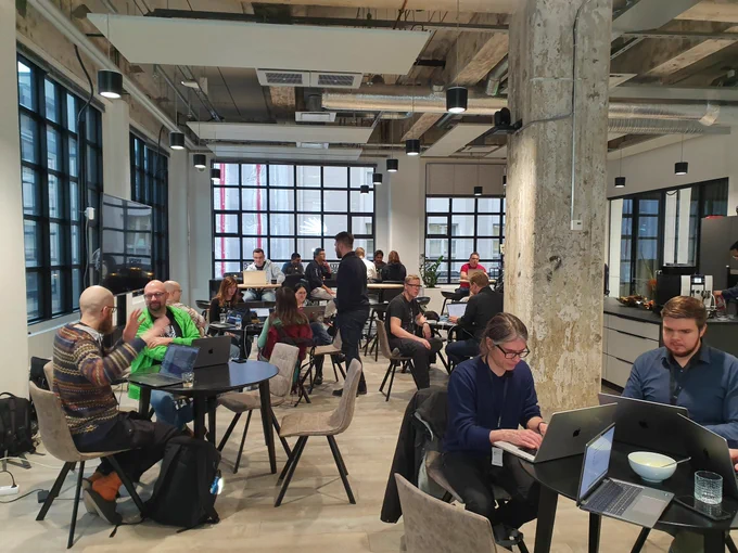 About 20 developers working on laptops in an office cafeteria. Lauri Eskola standing in the middle instructing a beginner contributor.