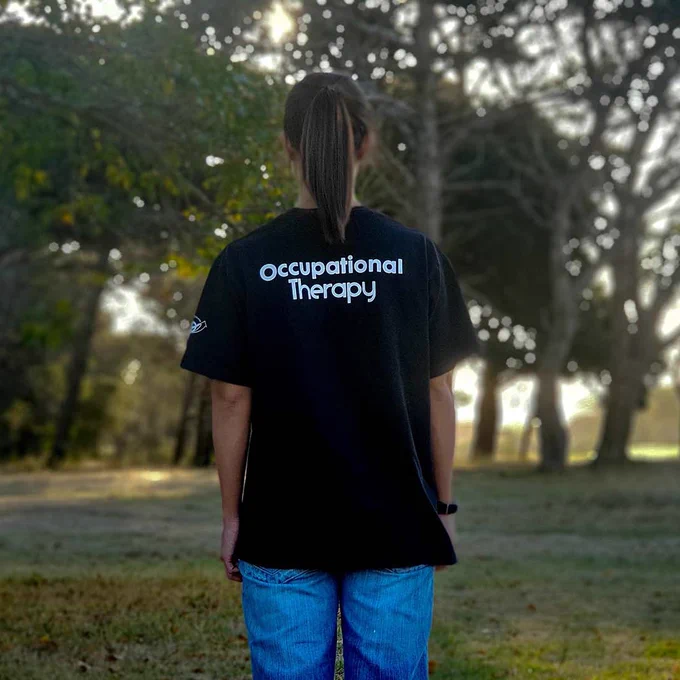 A person standing in a forest wearing a t-shirt that reads 'Occupational therapy', credit: Photo by Meelan Bawjee on Unsplash.