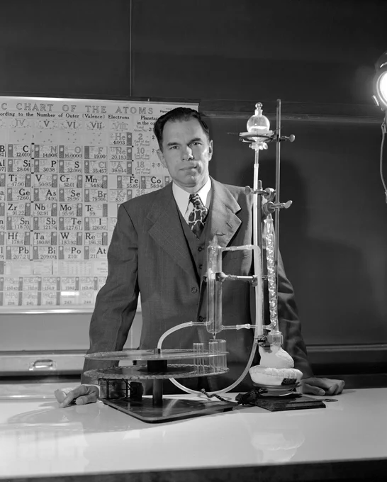 Seaborg standing in front of a period table. He is wearing a suit and a white shirt. 
