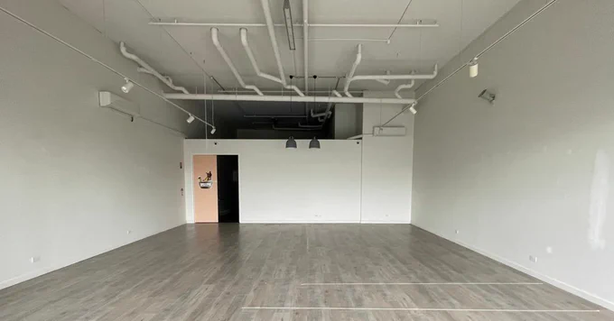  A photograph of an open space inside a building. The walls are white and on the ceiling, there  are visible pipes and lights. On the back door there is a door that is open. The floor has grey-ish floorboards.