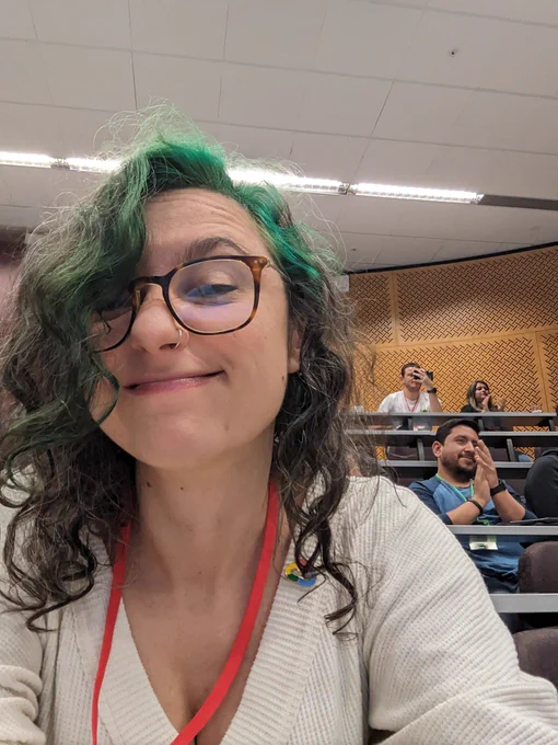 Photo of my face, in a lecture theatre. I'm squishing my face up a bit. I have dark curly hair with a green streak at one temple, and olive skin. I'm wearing a white sweater and a red lanyard 