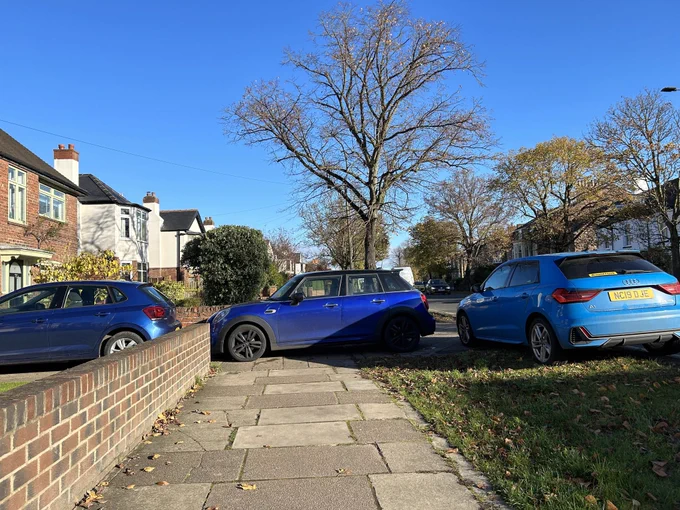 Three blue cars parked in a drive, across the pavement and on the verge without any gaps between them blocking the route entirely.