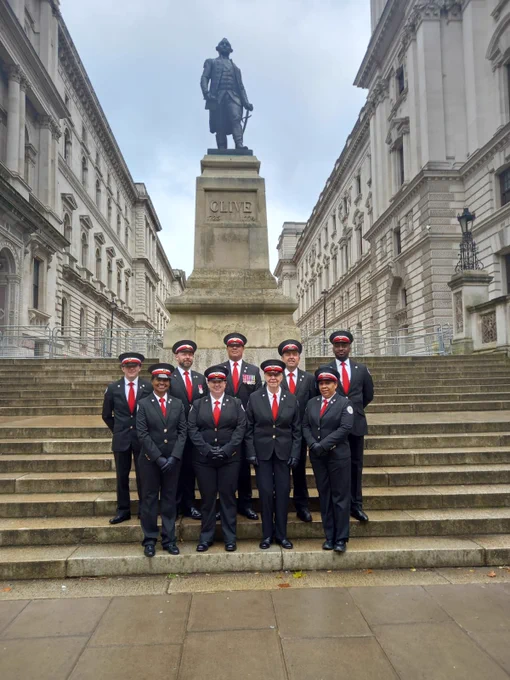 9 volunteers wearing standing in two rows at the Cenotaph in London. They are wearing red white and grey ceremonial uniform.