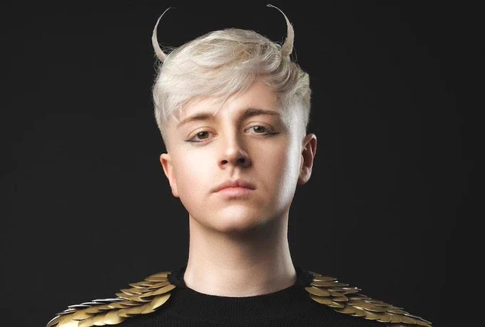 Photo of Daniel Foxx wearing a black top with gold metal discs covering the shoulders, and two strands of his hair styled upwards to resemble devil horns.