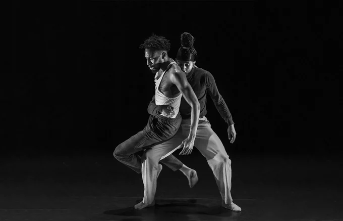 Black and white photo of two dance performers from FUBUNATION's 'RUINS'. They are wearing loose fitting clothing and are photographed mid-performance, one dancer has their legs apart with knees bent, and one arm around the waist of the other dancer holding them close their body. 