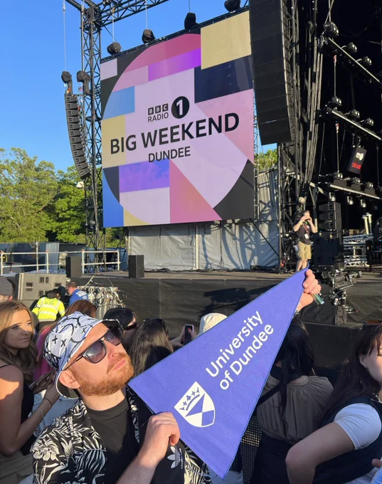 A man wearing a bucket hat and sunglasses holds a University of Dundee pennant flag in front of the stage at the BBC Radio 1 Big Weekend. A Big Weekend graphic is seen on the screen in the background