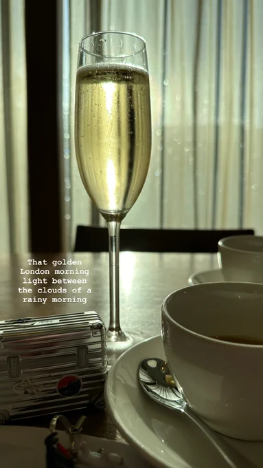 A glass of Champagne in the morning light on a table with a teacup and a small Rimowa Pilot Trolley style AirPods Pro holder. Caption: "that golden London mornign light between the clouds of a rainy morning".